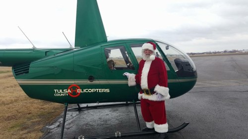 Santa Claus in a red suit stands next to a green helicopter with "Tulsa Helicopters" logo on a cloudy day.