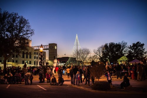 Crowd gathered at dusk for a festive event, featuring a lit Christmas tree and buildings in the background.