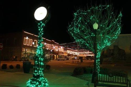 A clock tower adorned with green lights, surrounded by festive decorations and illuminated buildings at night.