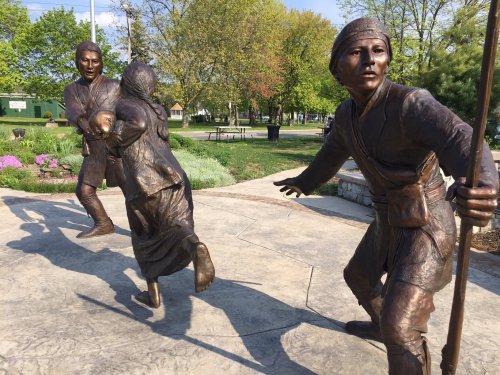 Bronze sculptures of three figures in a park, depicting movement and interaction among them. Green trees in the background.