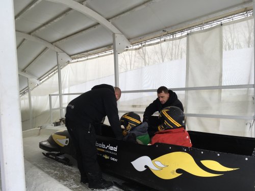 Two men assist two people in helmets inside a bobsled at a training facility.