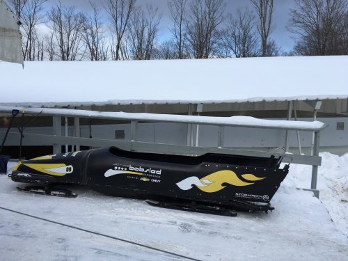A black bobsled with yellow accents parked on a snowy track, under a covered area surrounded by trees.