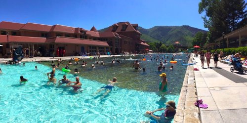 A busy outdoor pool scene with people swimming and relaxing, surrounded by mountains and sunny skies.