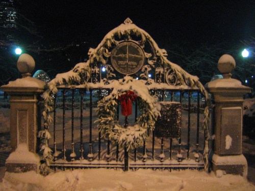 A snow-covered gate adorned with a wreath and red bow, illuminated by soft lights at night.