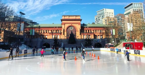 People ice skating on a rink in front of a historic building under a clear blue sky.