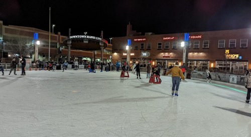 A lively outdoor ice skating rink at night, with people skating and enjoying the atmosphere in downtown Rapid City.
