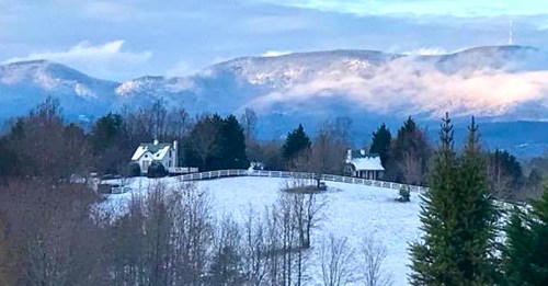 A snowy landscape with two houses, surrounded by trees and mountains under a cloudy sky.
