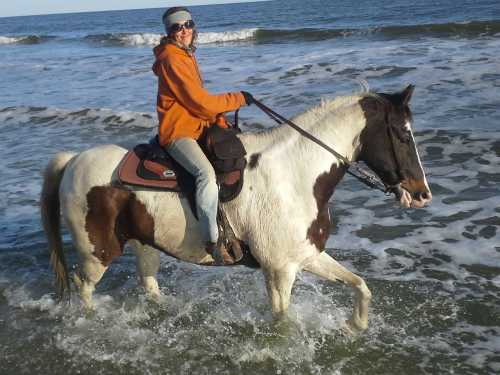 A person in an orange jacket rides a horse through shallow ocean water, with waves in the background.