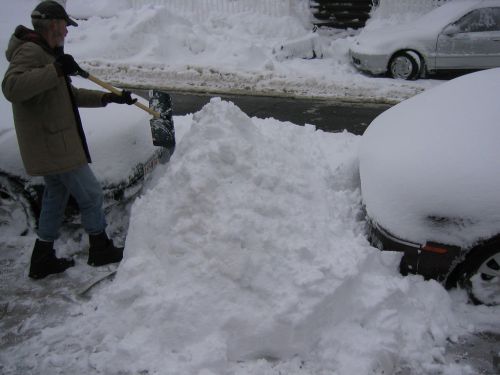 A person shovels snow from a street, with two cars partially buried under a thick layer of snow.