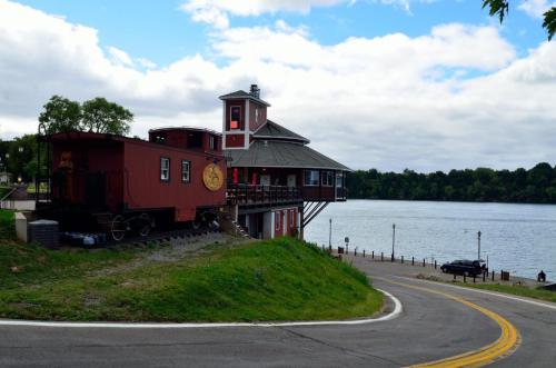 A red train car converted into a building near a river, with a winding road and green trees in the background.