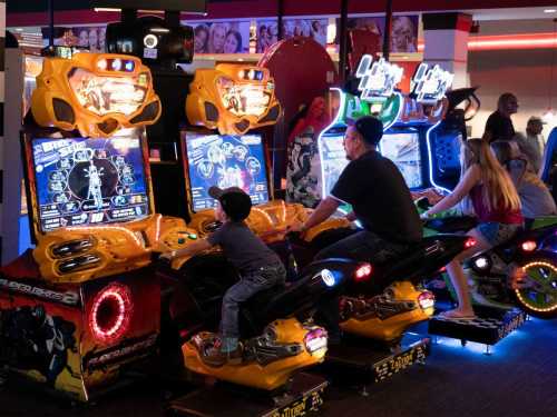 A child and an adult play racing arcade games in a vibrant arcade filled with colorful machines.