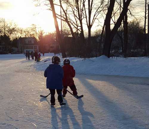 Two children skate on an outdoor ice rink at sunset, with trees and houses in the background.