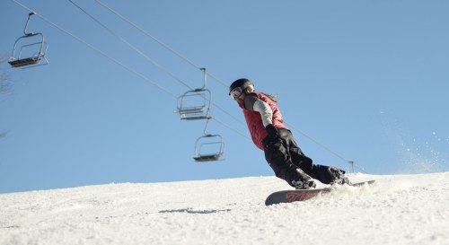 A snowboarder glides down a snowy slope under a clear blue sky, with ski lifts in the background.