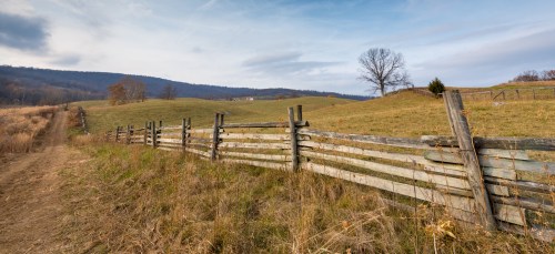A rustic wooden fence lines a grassy field, with rolling hills and a tree in the background under a cloudy sky.