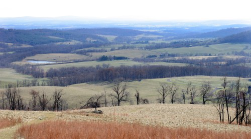 A panoramic view of rolling hills and fields, with sparse trees and a distant lake under a cloudy sky.