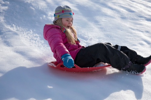 A child in a pink jacket and gray hat sleds down a snowy hill, enjoying a winter day.