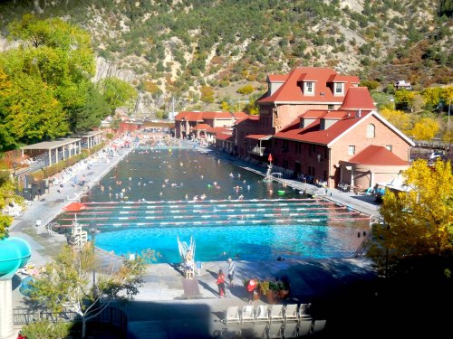 A large outdoor swimming pool surrounded by mountains, with people swimming and lounging on a sunny day.