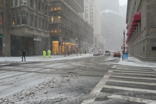 A snowy city street with pedestrians and vehicles, buildings partially obscured by falling snow.