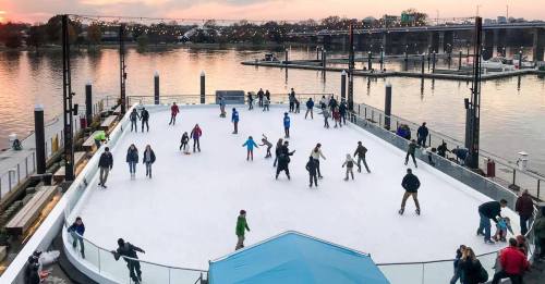 A bustling outdoor ice skating rink with people skating, surrounded by water and boats at sunset.