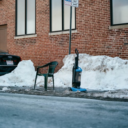 An empty chair and a vacuum cleaner sit on a snowy sidewalk next to a parked car and a brick building.