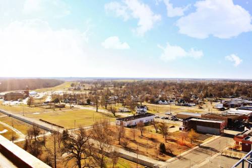 A panoramic view of a small town with open fields, roads, and scattered buildings under a bright blue sky.