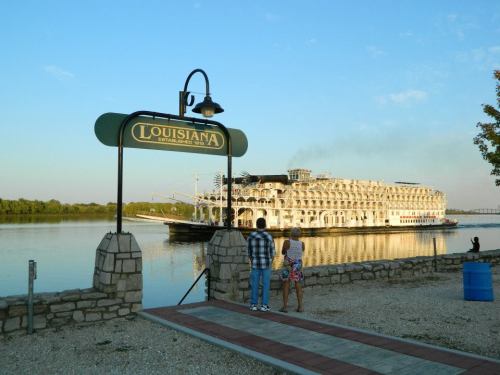 A couple stands by a sign for Louisiana, watching a large riverboat on the water under a clear blue sky.