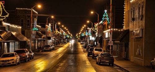 A quiet street at night, lined with festive holiday lights and decorated trees, reflecting on wet pavement.