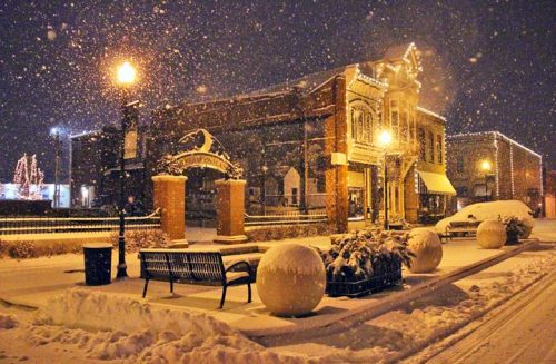 A snowy night scene of a quaint town square, featuring a decorated building, benches, and large snow-covered spheres.