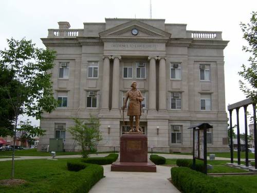 Statue of a man in front of a grand, historic building with columns and a clock, surrounded by greenery.