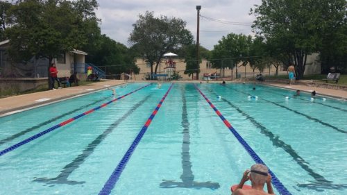 A clear swimming pool with lanes, surrounded by trees and people enjoying the area on a cloudy day.
