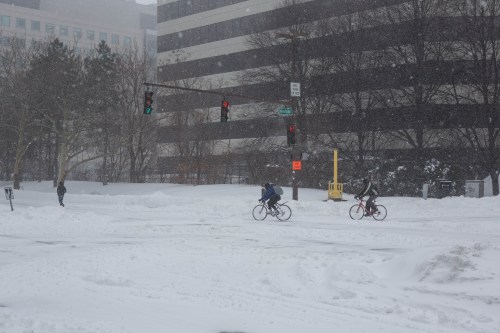 Two cyclists navigate a snowy street while a pedestrian waits at a traffic light in a winter storm.