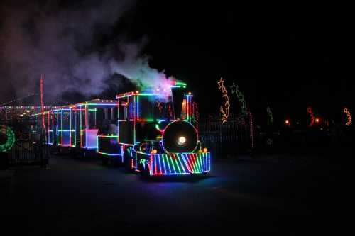 A brightly lit train decorated with colorful lights, emitting steam against a dark night sky.