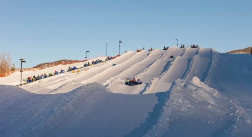 A snowy hill with people sledding down on tubes, surrounded by trees and clear blue skies.