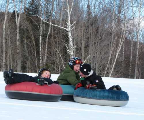 Three children sit on colorful snow tubes, smiling and enjoying a winter day in a snowy landscape with trees in the background.