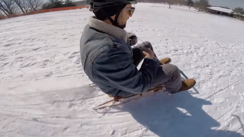 A person in a denim jacket and helmet sledding down a snowy hill, holding a camera.