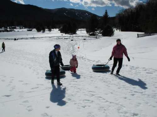 Three people pulling snow tubes across a snowy landscape with mountains in the background on a sunny day.