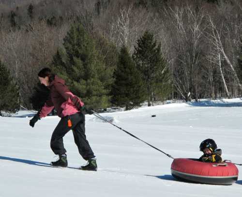 A person pulls a child on a snow tube through a snowy landscape, surrounded by trees.