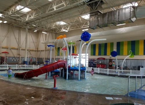 Indoor water play area featuring slides, water features, and colorful umbrellas. Children are playing in the shallow water.