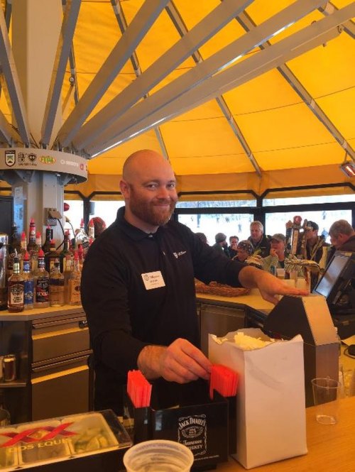 A smiling bartender stands behind a bar, preparing drinks with a lively crowd in the background under a yellow canopy.
