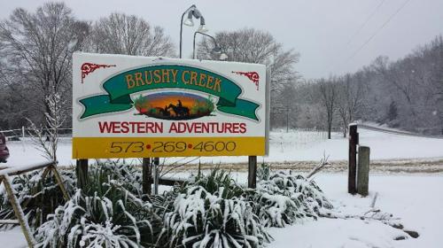 Sign for Brushy Creek Western Adventures covered in snow, with trees and a snowy road in the background.