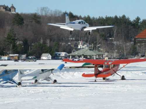 A small plane is landing over a snowy airfield with other aircraft parked nearby and trees in the background.