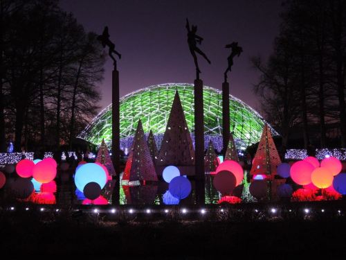 Colorful illuminated decorations and trees in a park at night, with a large green dome structure in the background.