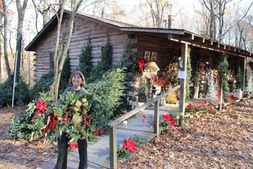 A woman stands outside a rustic log cabin decorated with greenery and red bows, surrounded by autumn leaves.
