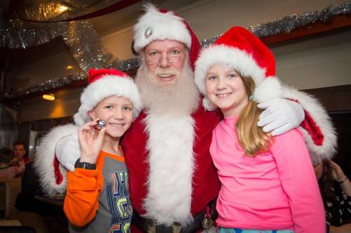 Santa Claus poses with two children, one holding a small toy, both wearing festive hats and smiling joyfully.