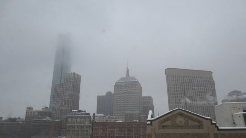A snowy city skyline shrouded in fog, featuring tall buildings and a gray sky.