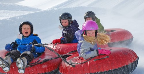 Four children joyfully tubing down a snowy slope, wearing helmets and colorful winter gear, with snow flying around them.