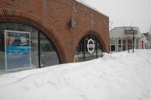 A brick building with a lottery sign, partially buried in snow, next to a storefront in a snowy urban setting.