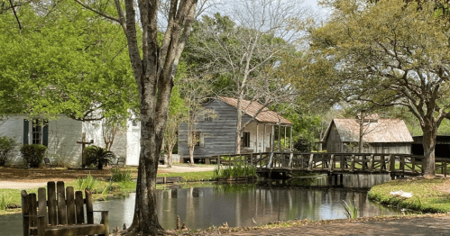 A serene scene of a pond surrounded by trees, with rustic buildings and a wooden bridge in a peaceful setting.