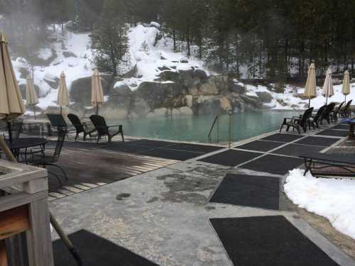 A serene hot spring pool surrounded by snow, with lounge chairs and umbrellas, set against a rocky backdrop.