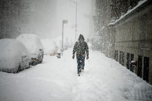 A person walks through a snowy street, with cars buried in snow and heavy snowfall obscuring visibility.